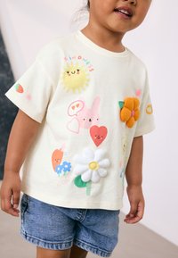 Cream short-sleeve t-shirt featuring colorful, embroidered appliqués of flowers, sun, and hearts, with denim shorts.