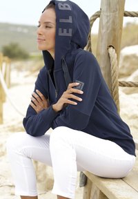 Felpa navy con zip frontale, grande cappuccio e stampa del logo. Abbinata a pantaloni bianchi a vita alta. La texture appare morbida e liscia.