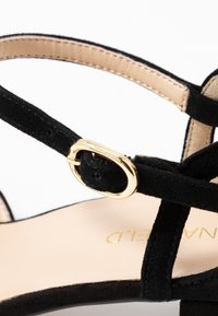 Sandal de ante negro con hebilla dorada, diseño de tiras entrecruzadas y plantilla beige con el nombre de la marca.