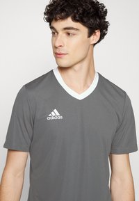 Camisa atlética gris de manga corta con escote en V, cuello blanco y un logo de Adidas en blanco en el lado izquierdo. Textura de tela suave.