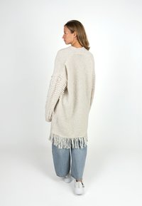 Frau steht und schaut zur Seite, trägt eine lange, cremefarbene, fransige Strickjacke, weite, blaue Jeans und weiße Sneakers.