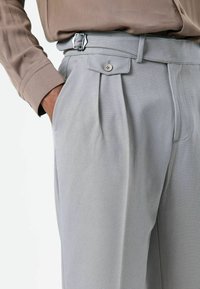 Pantalon gris clair tailleur avec plis, passants pour ceinture et fermeture par bouton, fabriqué en tissu texturé. Poche latérale visible.