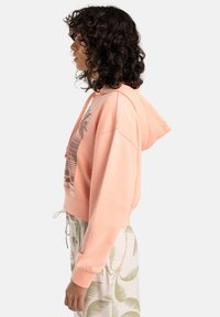 Perzikkleurige cropped hoodie met een palmboomgrafiek, voorzien van een capuchon met trekkoord, geribbelde manchetten en een relaxed fit, gecombineerd met lichtgroene broek met palmenpatroon.