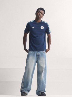 Młody mężczyzna stojący z rękami w kieszeniach, ubrany w granatowy t-shirt Adidas z jasnoniebieskimi paskami oraz szerokie jasnoniebieskie dżinsy na jednolitym tle.