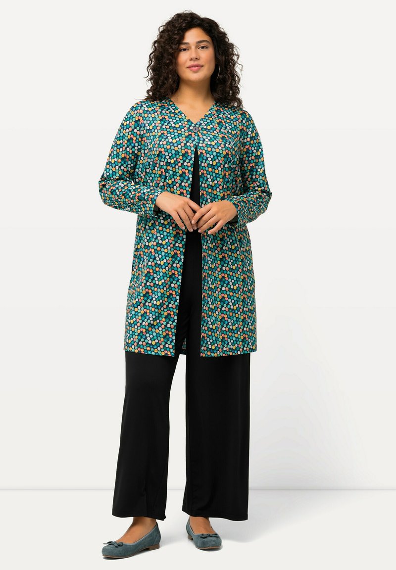 Lungo cappotto turchese con un motivo a pois colorati, abbinato a pantaloni neri a gamba larga e scarpe piatte teal. Presenta un collo a V e chiusura con bottoni.