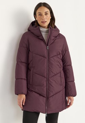 Woman wearing a knee-length, purple puffer coat with a hood, standing against a plain light background.