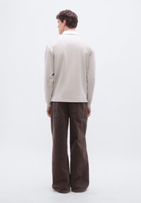 Bărbat stând cu spatele, purtând o cămașă bej cu mâneci lungi și pantaloni largi maro închis, pe un fundal alb.