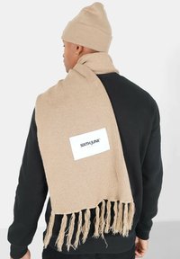 Beige gebreide sjaal met franje-einden en een wit genaaid label met de tekst "SIXTH JUNE." Draag over een zwarte sweatshirt met een ronde hals.