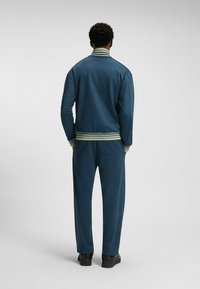Herre tracksuit i m�ørk teal med kontrasterende grønne striber på kraven og ærmerne. Den har en afslappet pasform og en glat stofstruktur.