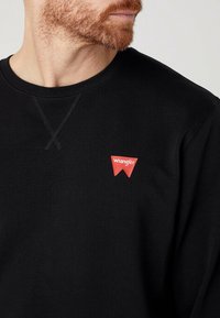 Svart sweatshirt med en röd triangulär "Wrangler"-logotyp på vänstra bröstet. Har en ribbad krage och texturerad söm.