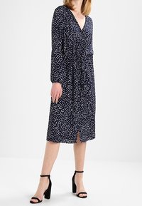 Abito blu navy con pois bianchi, scollatura a V, maniche lunghe, vita aderente e orlo al ginocchio. Indossato con sandali neri con tacco.