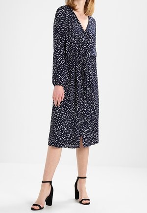 Robe bleu marine avec des pois blancs, décolleté en v, manches longues, taille cintrée, et ourlet jusqu'aux genoux. Portée avec des sandales à talons noires.