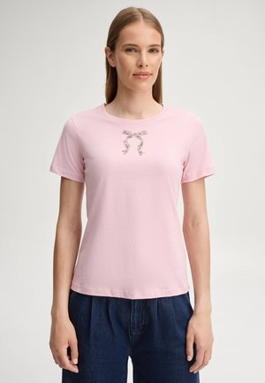 Vrouw met steil lichtbruin haar draagt een lichtroze t-shirt met een kleine strikprint en donkerblauwe broek, staand tegen een effen achtergrond.