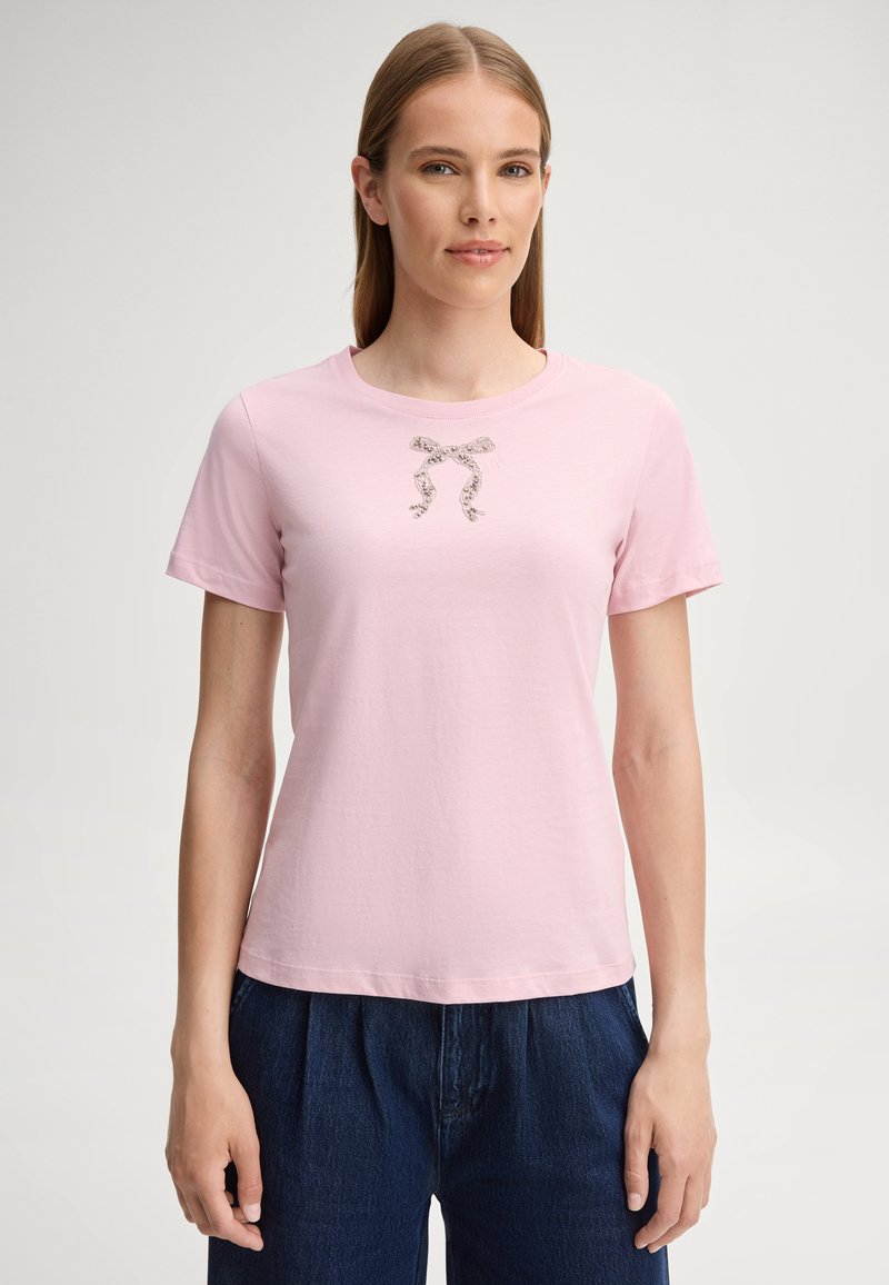 Frau mit glattem, hellbraunem Haar trägt ein hellrosa T-Shirt mit einer kleinen Schleifengrafik und dunkelblaue Hose und steht vor einem schlichten Hintergrund.