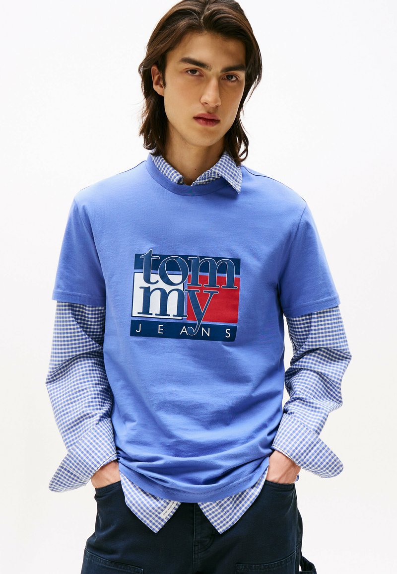 Joven con cabello largo que lleva una camiseta azul de Tommy Jeans sobre una camisa de cuadros azul de manga larga, con las manos en los bolsillos de unos pantalones oscuros, fondo neutro.