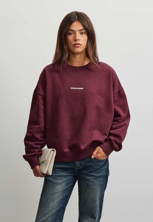 Ung kvinde med langt brunt hår iført en vinrød sweatshirt med "PEGADOR" tekst, blå jeans og holder en sammenfoldet lys beige clutchtaske.