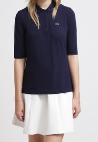 Mörkblå pikétröja med korta ärmar, treknappsstängning och Lacoste-logotyp broderad på bröstet, matchad med en strukturerad vit kjol.