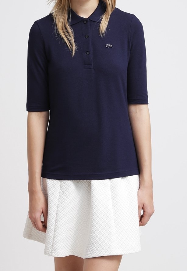 Mörkblå pikétröja med korta ärmar, treknappsstängning och Lacoste-logotyp broderad på bröstet, matchad med en strukturerad vit kjol.