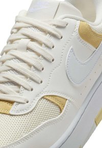 Zapatillas de piel blanca con acentos de malla, detalles dorados y un swoosh blanco contrastante. Presenta cordones planos y lengüeta acolchada.