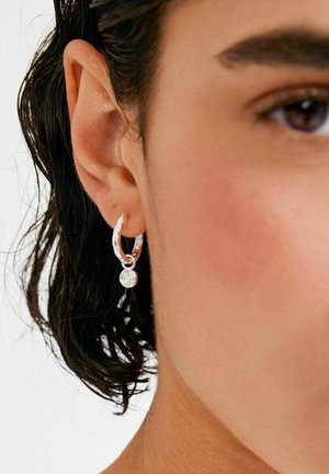 Pendientes de aro plateados con un diseño trenzado y un pequeño colgante circular con un detalle texturizado. Se llevan en la oreja.