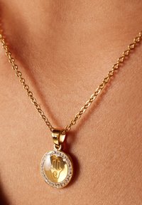 Gouden ketting met een ronde hanger met een hand symbool, omringd door heldere stenen, hangend aan een schakelketting.