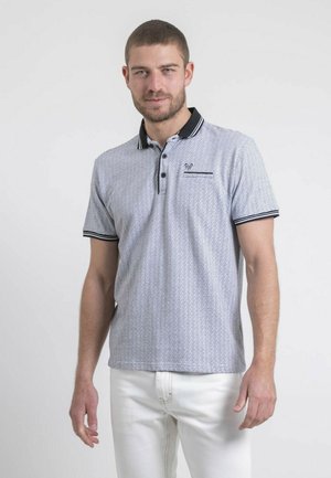 Homme portant un polo gris clair à motifs avec des bordures noires et un pantalon blanc, debout devant un fond clair uni.
