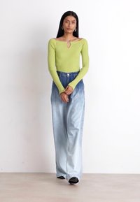 Top lungo a maniche lunghe verde a coste con dettaglio a chiave, abbinato a jeans a gamba larga azzurri. Scarpe nere completano l'outfit.