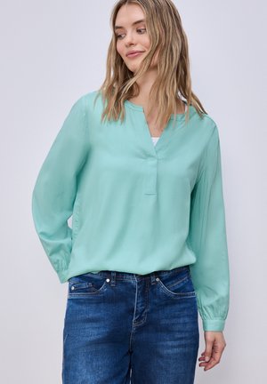 Vrouw die een licht turquoise blouse met lange mouwen en een blauwe spijkerbroek draagt, lichtjes opzij kijkt met een zachte glimlach.