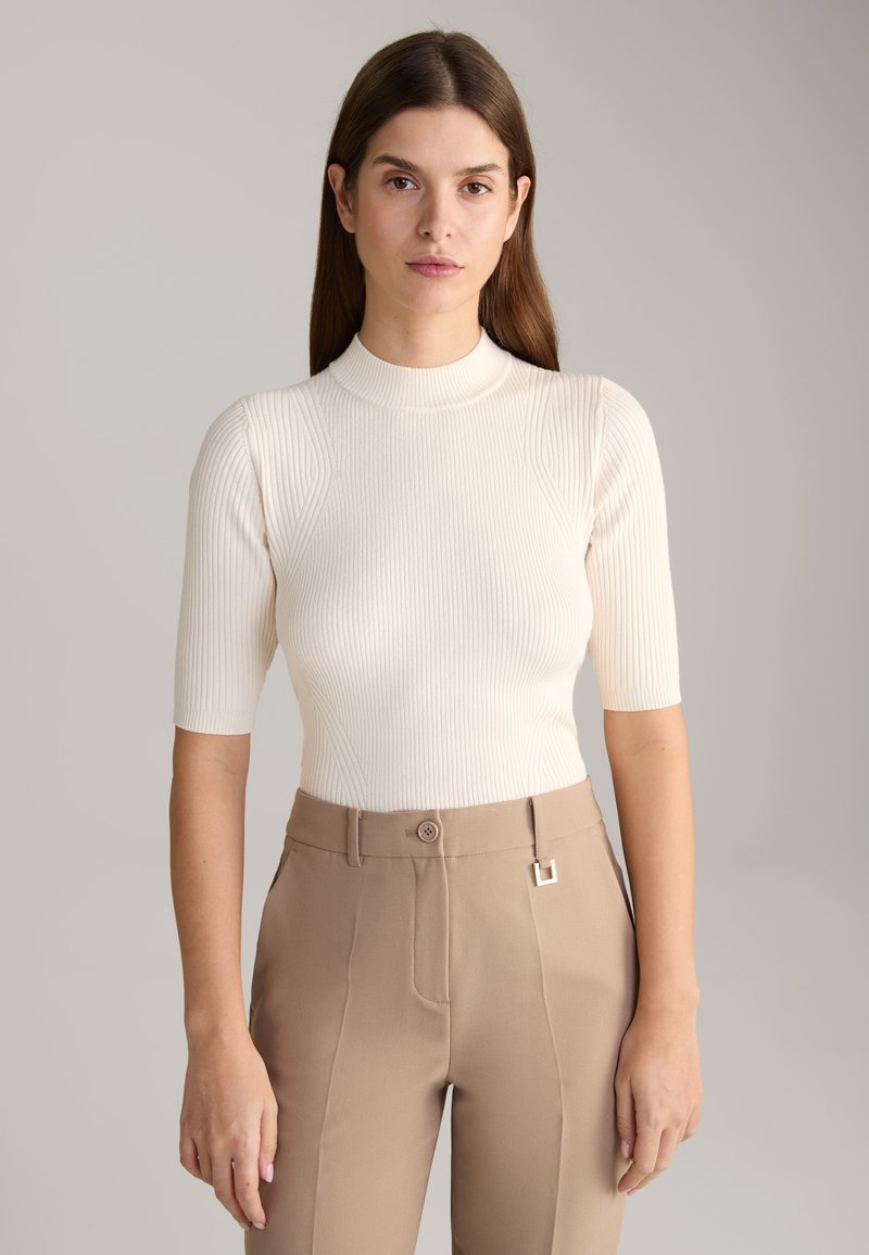 Haut en maille côtelée beige clair avec un col haut et des manches courtes, associé à un pantalon tailleur taupe doté d'un bouton et de quincaillerie décorative.