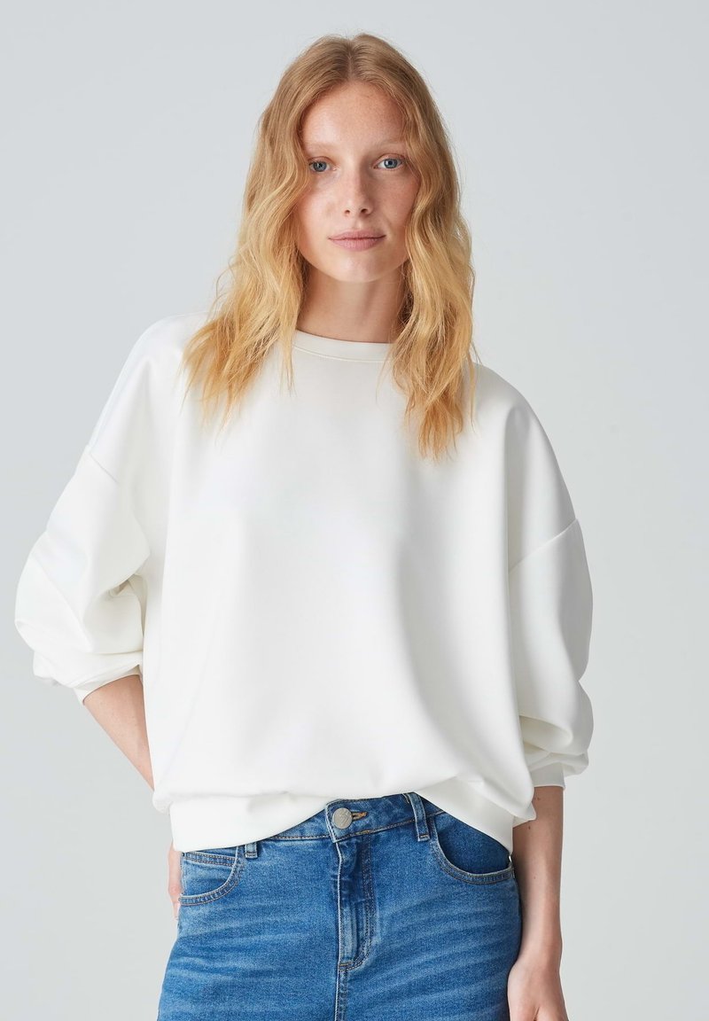 Sweatshirt bianco oversize con scollatura rotonda, spalle scese e polsini a costine, abbinato a jeans fitted blu chiaro.