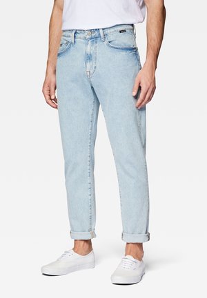 Mand iført lyseblå opkrogede jeans, hvide sneakers og en hvid t-shirt med afslappede hænder langs siden.