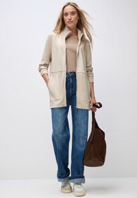 Cardigan beige à fermeture éclair avec col montant, deux poches avant, associé à un jean large bleu clair et des baskets blanches. Sac à main en daim marron.