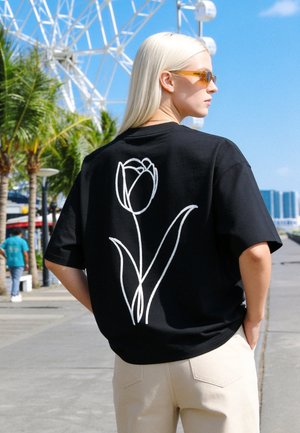 Femme blonde portant des lunettes de soleil et un t-shirt noir avec un croquis blanc de tulipe marche le long d'une promenade ensoleillée avec des palmiers et une grande roue.
