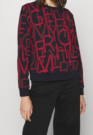 Persoon draagt een marineblauwe trui met overlappende rode letters en zwarte broek, staand tegen een effen lichte achtergrond.