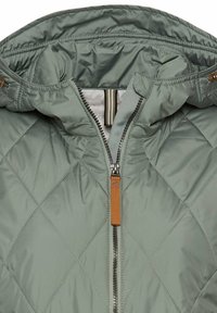 Veste matelassée vert clair avec un col montant et une fermeture éclair avant avec une tirette orange et une doublure intérieure rayée.