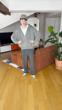 Nagyméretű szürke cipzáras kapucnis pulóver fehér inggel, szürke nadrággal és ezüstös tornacipővel párosítva. Beltéri környezetben, fa padlóval és növényekkel.