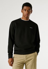 Sweatshirt preto com decote redondo e mangas compridas. Apresenta um pequeno logotipo branco no peito e uma bainha com efeito canelado. Estilizado com calças de veludo cotelê bege.