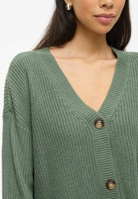 Cardigan vert tricoté avec un col en V, présentant un motif texturé et deux boutons marron à l'avant. Tissu doux avec une coupe décontractée.