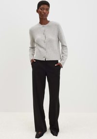 Vrouw in een lichtgrijze gebuttonde cardigan, zwarte wijde broek en zwarte puntige schoenen, met haar handen in de zakken.