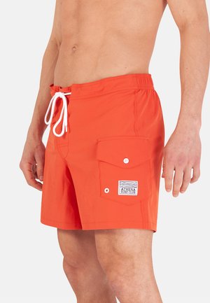 Torse masculin portant un short de bain orange vif avec un cordon blanc et une poche latérale étiquetée « Athena Surf Club » sur fond gris.