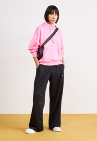 Maglione rosa con tasca, pantaloni neri larghi, scarpe bianche slip-on e una piccola borsa a tracolla nera. Texture morbide e vestibilità rilassata.