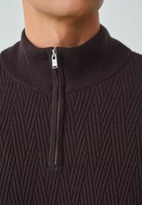 Donkerbruine zip-up trui met hoge kraag, voorzien van een gestructureerd zigzagpatroon en een zilveren ritsaccent bij de hals.