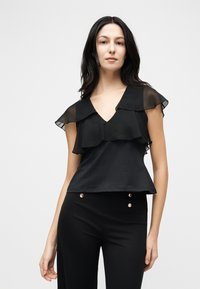 Γυναίκα με μακριά σκούρα μαλλιά που φοράει μαύρο αμάνικο τοπ με διαφανείς βολάν και μαύρο ψηλόμεσο παντελόνι με χρυσά κουμπιά.