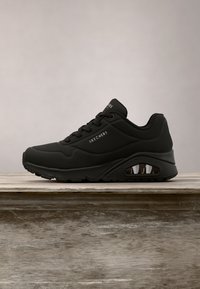 Baskets de sport noires Skechers avec détail visible de semelle à coussin d'air, design à lacets et logo de marque sur le côté et la languette, sur surface en bois.