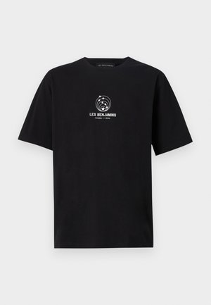 Henkilö, jolla on musta "Les Benjamins" T-paita, beige leveälahkeiset housut, valkoiset tennarit ja joka kantaa mustaa olkalaukkua.
