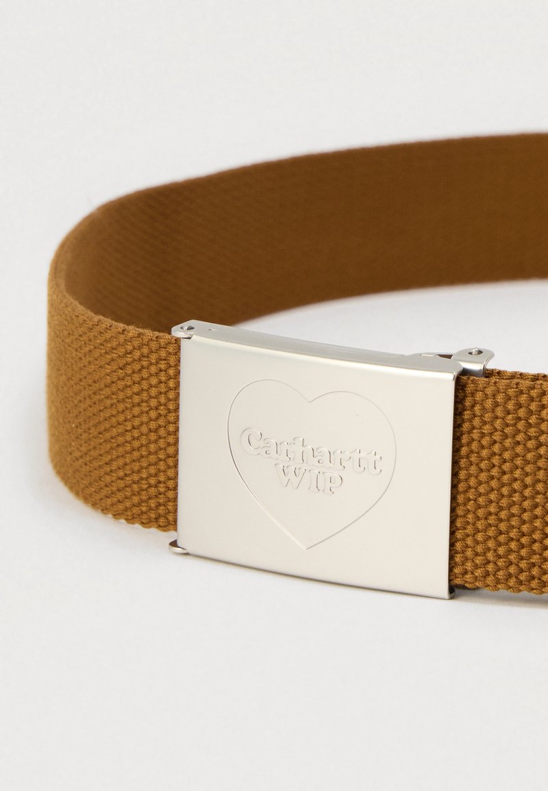 Cintura in fettuccia di cotone marrone con fibbia in metallo argento. La fibbia presenta un design a cuore in rilievo e il testo "Carhartt WIP."