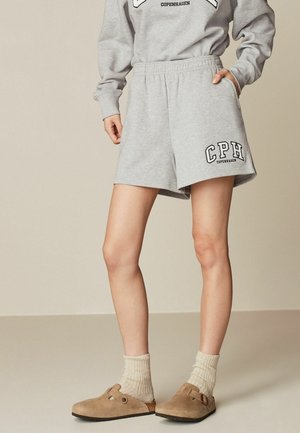 Personne portant un short gris clair avec le texte "CPH Copenhagen", des chaussettes beige et des chaussures à enfiler couleur tan sur un fond uni.