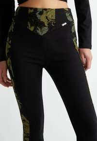 Leggings de cintura alta en negro con acentos florales verdes en los lados, con un patrón texturizado y un pequeño logo de la marca en la cintura.