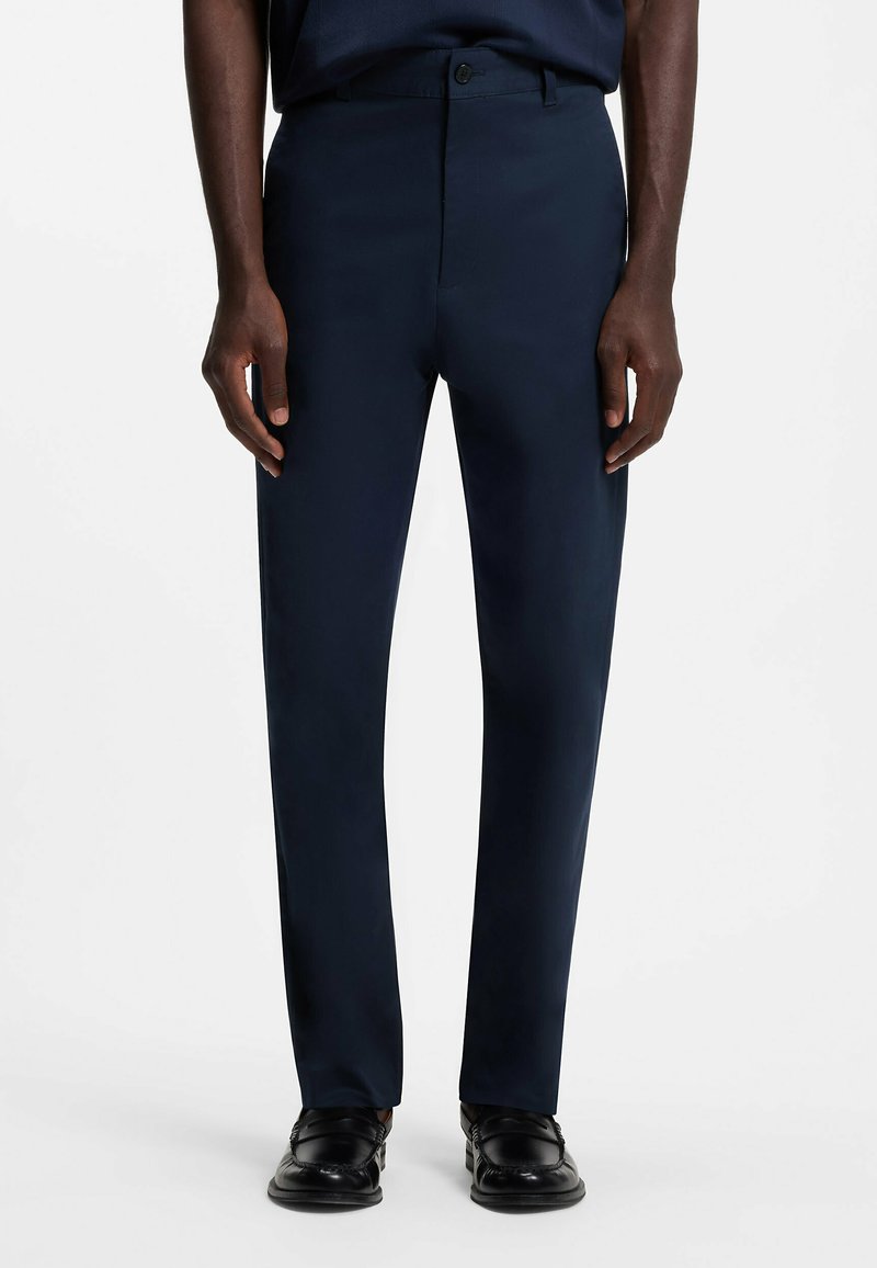 Homme portant un pantalon tailleur bleu marine foncé et des mocassins en cuir noir, debout sur fond blanc.