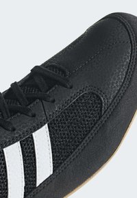 Gros plan d'une chaussure de sport noire avec des rayures blanches, présentant un tissu texturé et un matériau synthétique lisse avec des lacets noirs.
